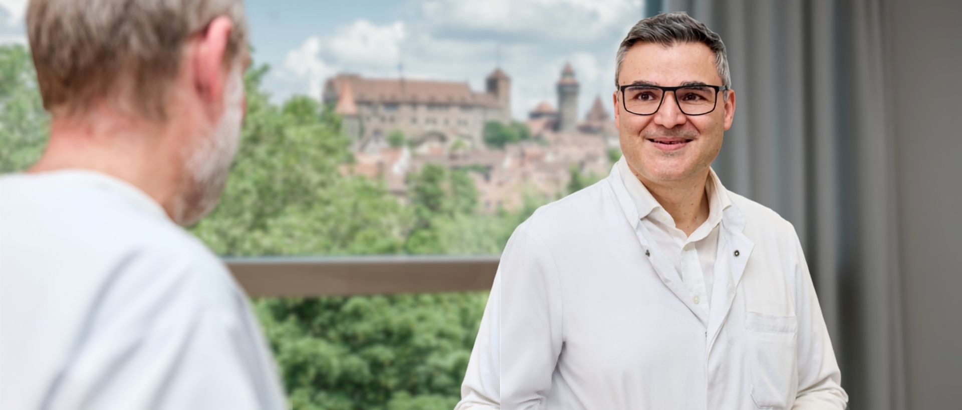 Dr. med. Dimitrios Vlachos, Leitender Arzt für Hüfterkrankungen in der Klinik für Orthopädie der DR. ERLER KLINIKEN, im Gespräch mit einem Patienten – im Hintergrund die Nürnberger Stadtsilhouette.