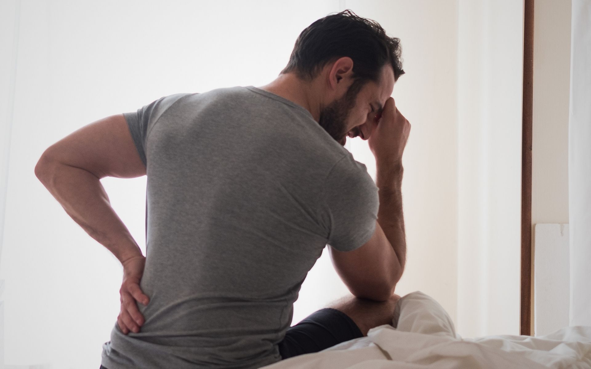 Ein Mann hält sich mit der einen Hand den Rücken, die andere Hand hat er in seinem Gesicht vergraben. Sein Gesichtsausdruck drückt aus, dass er unter Schmerzen leidet.