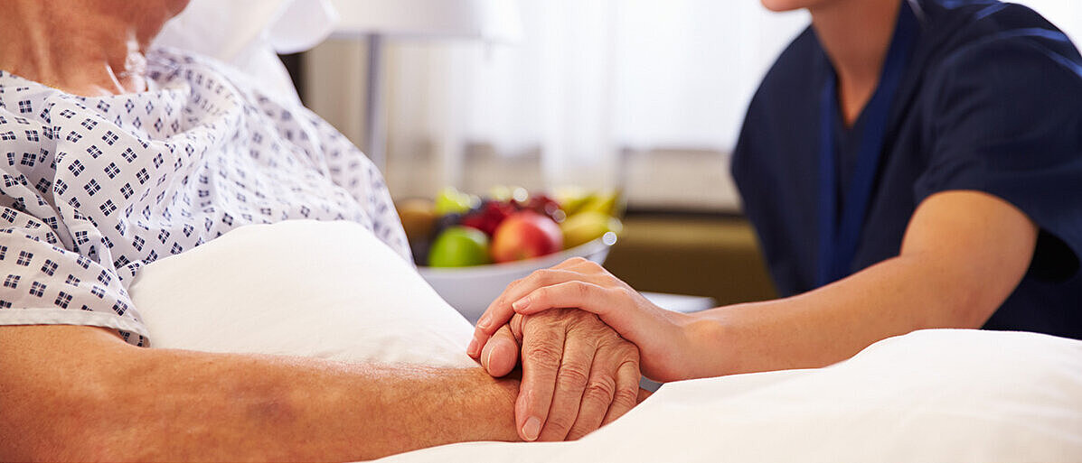 Eine Pflegekraft hält die Hand eines Patienten, der im Krankenbett liegt. 